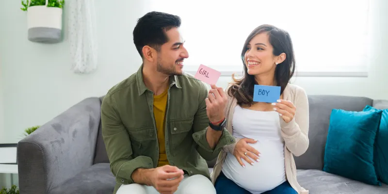 Expecting couple holding "girl" and "boy" cards on a couch.